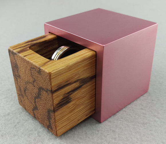 Pink metal engagement ring box, slightly open showing tan marble wood insert with black streaks