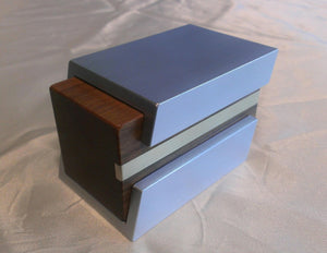 Silver metal engagement ring box with brown wood sliding insert. Metal shell has a cutaway to expose brass line inlay in wood